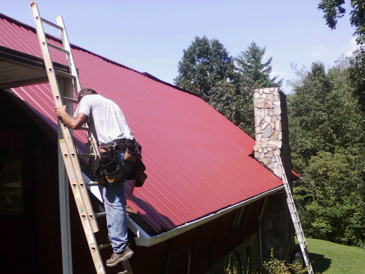 Roofer on a ladder beside a red metal roof and stone chimney — Soffit & Fascia Repair by Roofing Brothers in Jefferson City